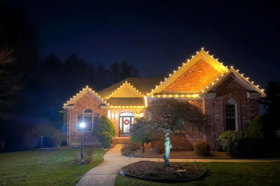 A brick ranch with warm white roofline lights and a small tree lit in the front bed beside the walkway.