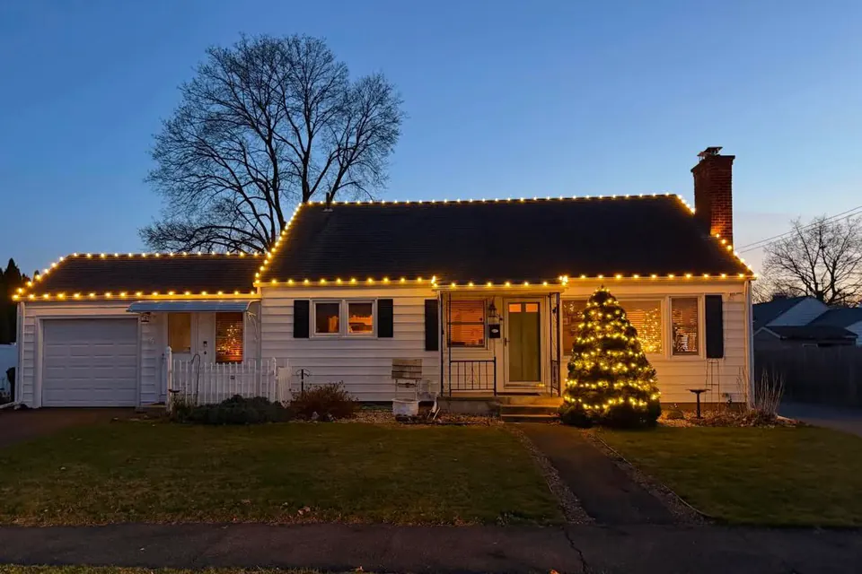 A cape with warm white roofline lights and a lit evergreen by the front door, photographed at dusk.
