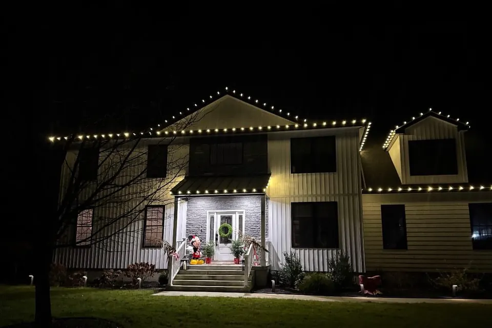 A two-story home with warm white roofline lights, a lit wreath on the front door, and uplights along the front steps.