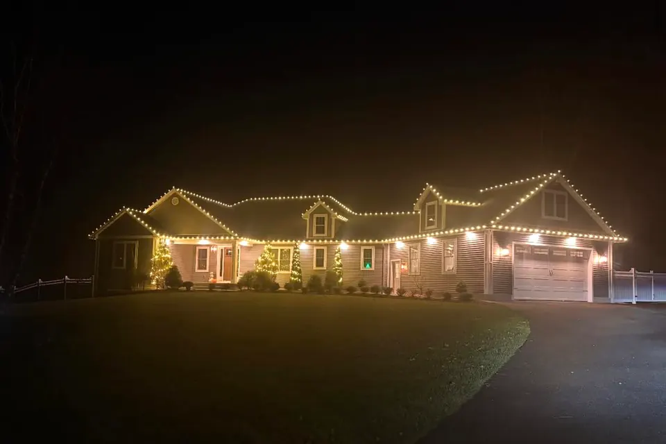 A long low home with warm white lights tracing several gable peaks, lit shrubs along the front, and a curving driveway.