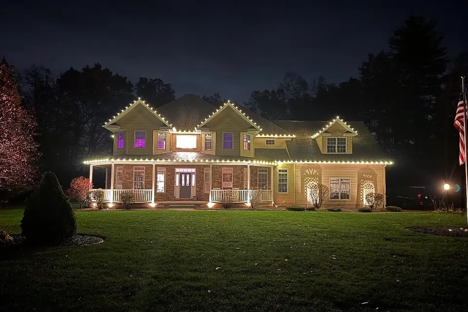A two-story home with a full front porch, warm white lights on the roofline and porch edge, and uplights on the columns.