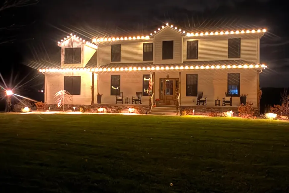 A two-story colonial with warm white lights along the roofline and the porch roof, rocking chairs on the porch, and ground lights washing the front.
