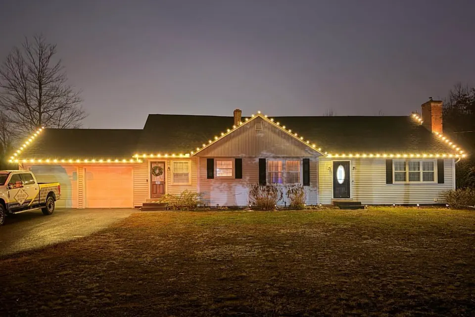 A ranch home with warm white roofline lights and a work truck in the driveway, photographed at dusk.