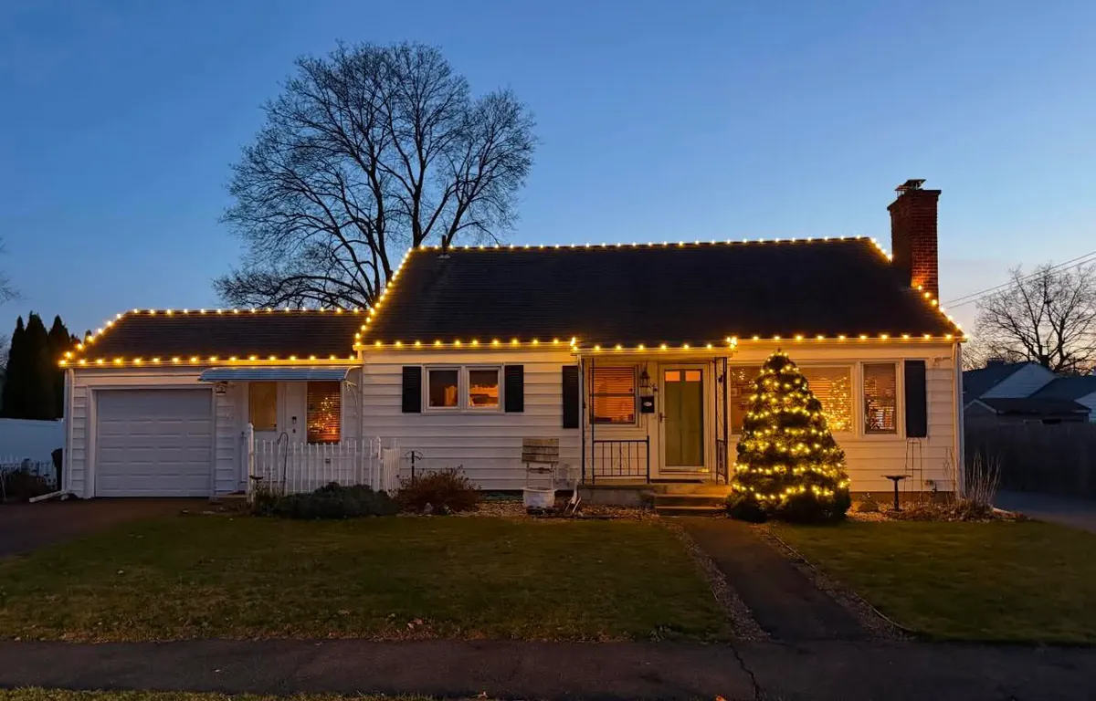 A cape with warm white roofline lights and a lit evergreen by the front door, photographed at dusk.