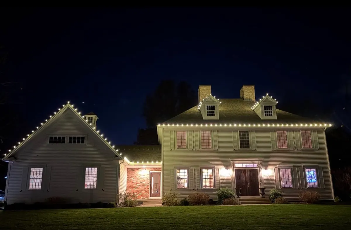 A white colonial with an attached wing, warm white lights along both rooflines and around the dormer windows.