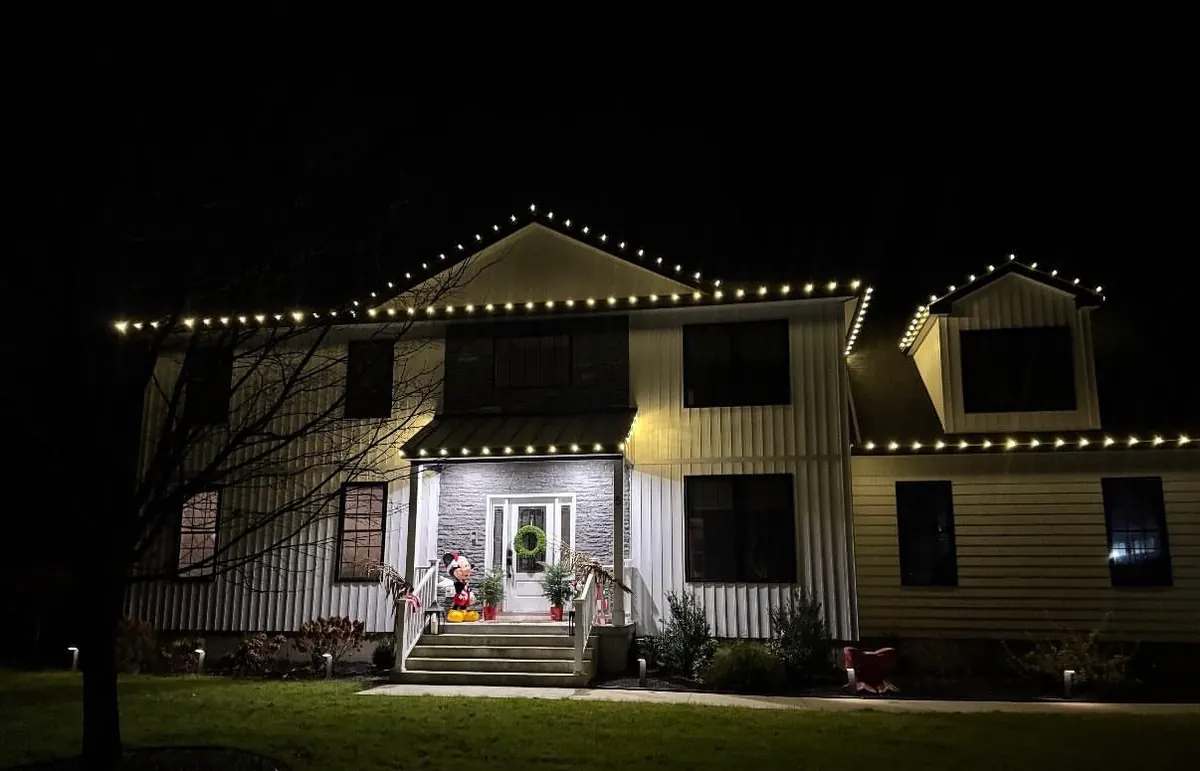 A two-story home with warm white roofline lights, a lit wreath on the front door, and uplights along the front steps.