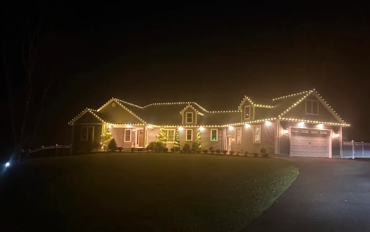 A long low home with warm white lights tracing several gable peaks and lit shrubs along the front.
