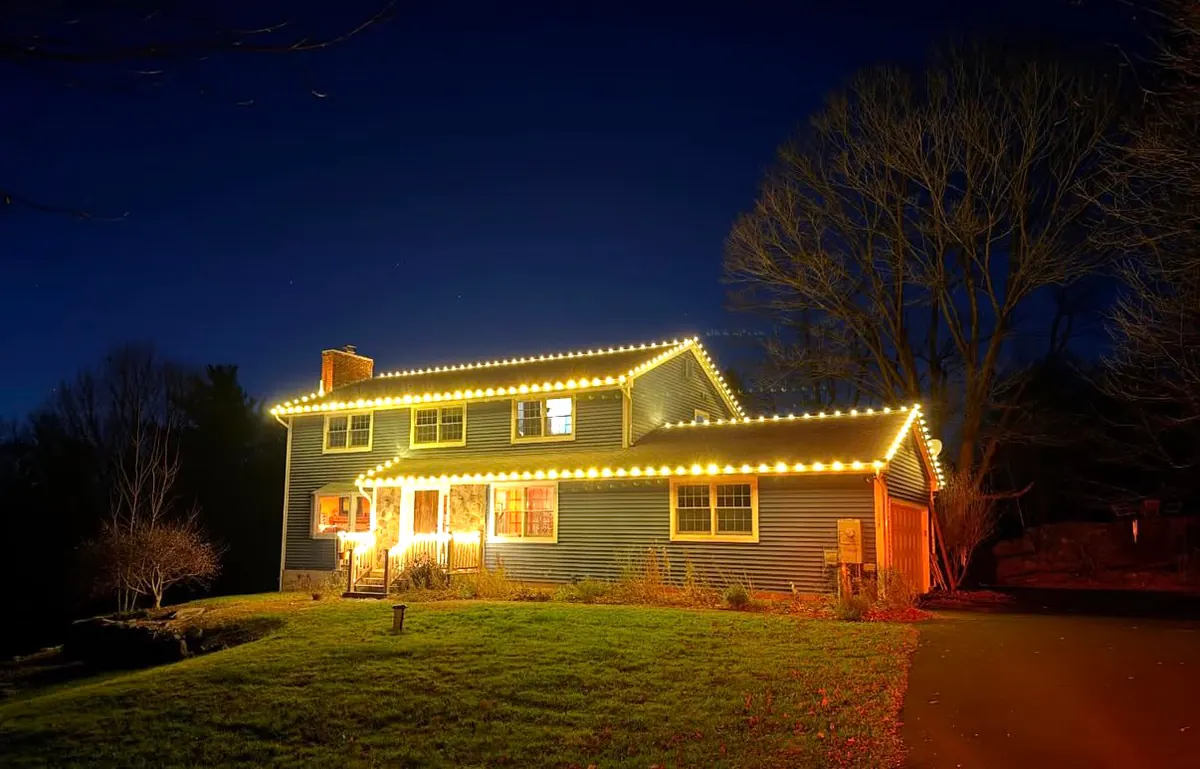 Warm white roofline lights on a two-story colonial against a deep blue dusk sky.