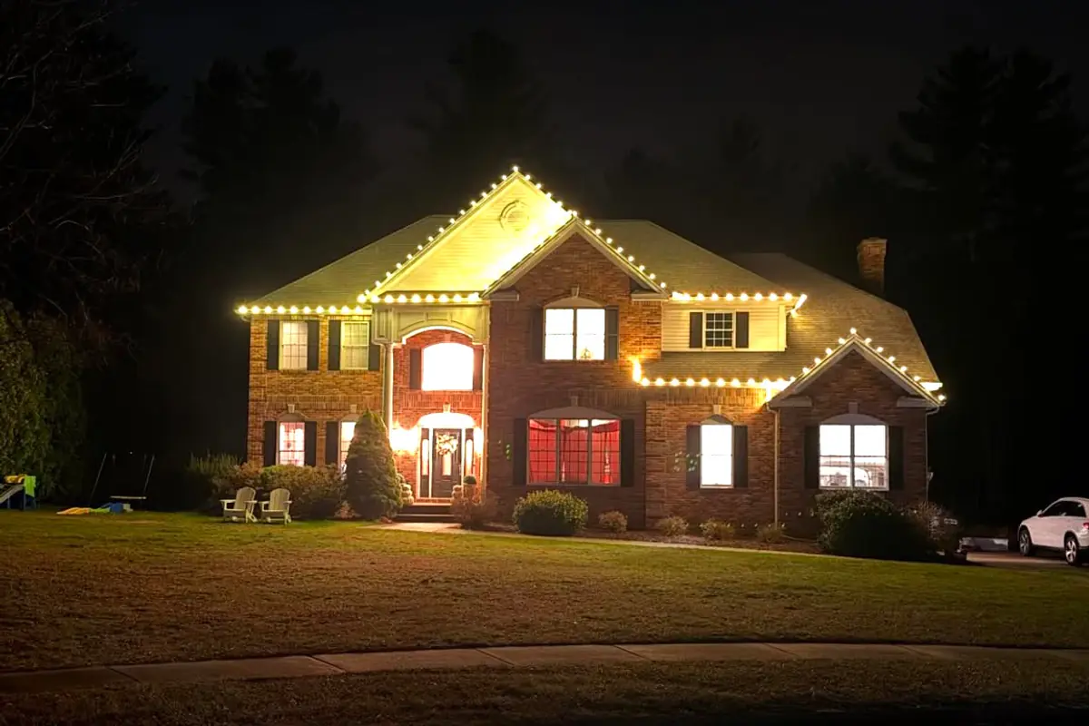 Two-story brick colonial with warm white lights along the roofline, gables and window trim on a clear evening.
