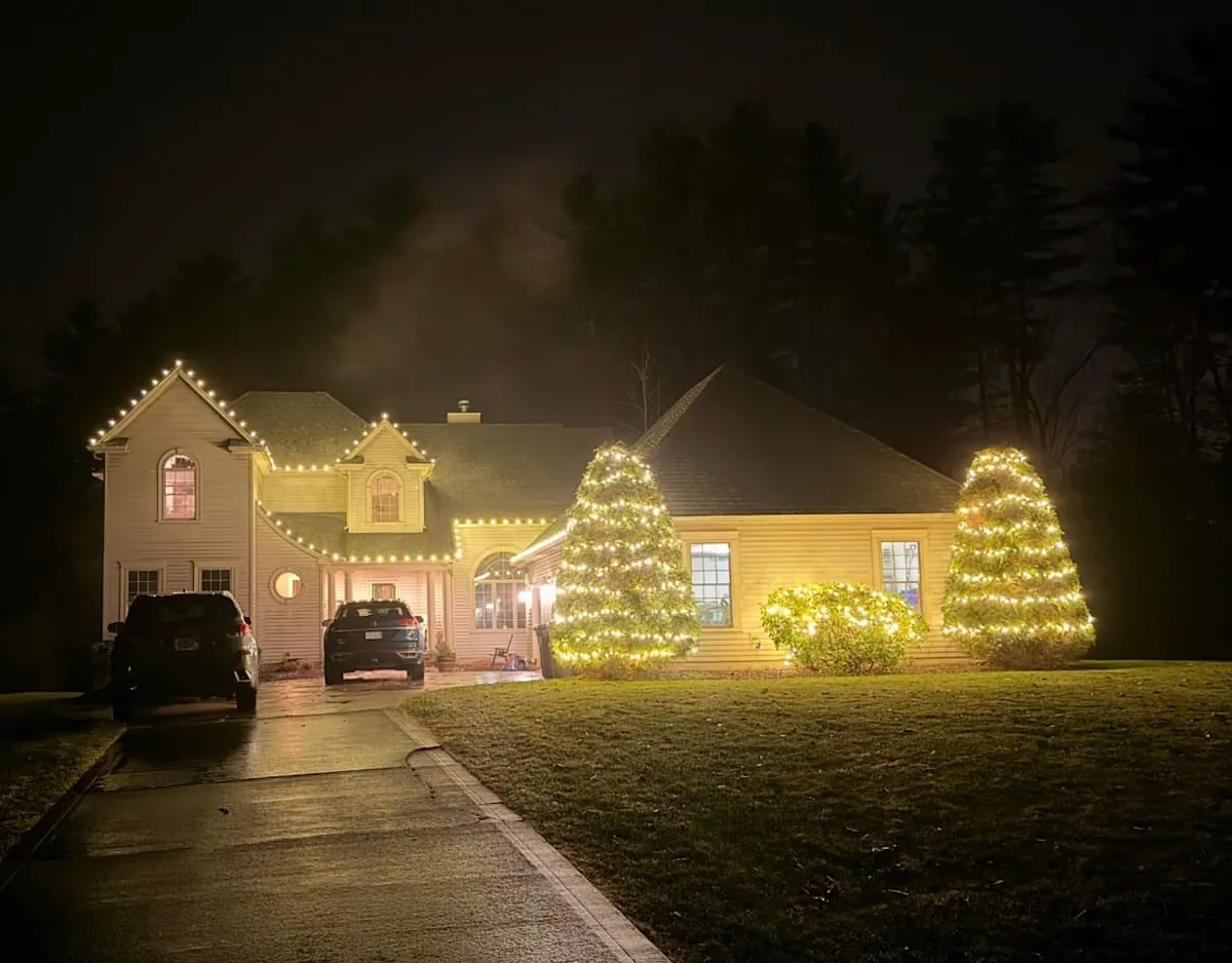 A two-story home at dusk with warm white roofline lights, two lit cone-shaped evergreens and a lit foundation bush across the front, with cars in the driveway.