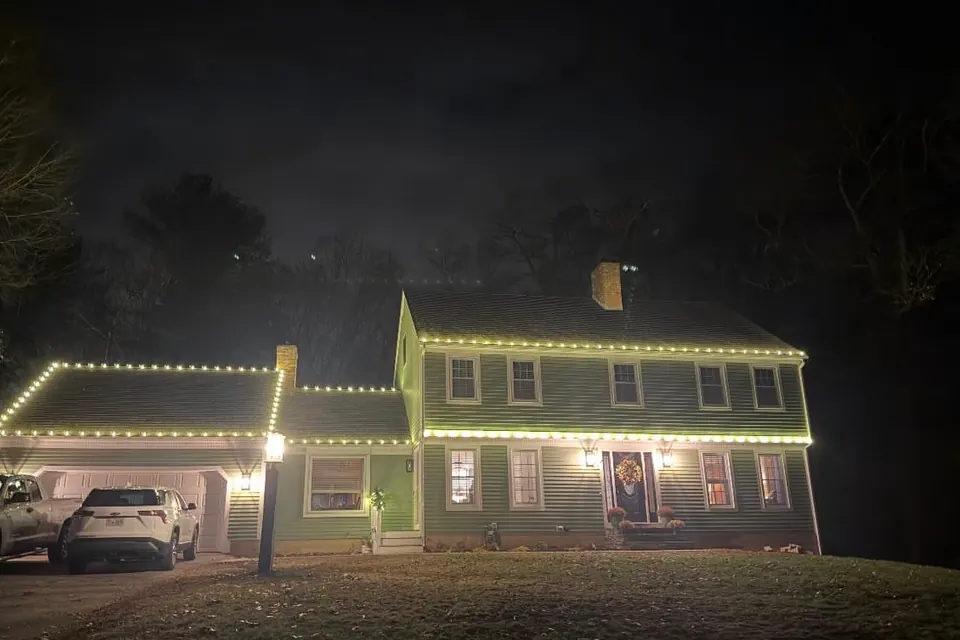 Warm white roofline lights on a two-story colonial photographed from the road across a deep front lawn.